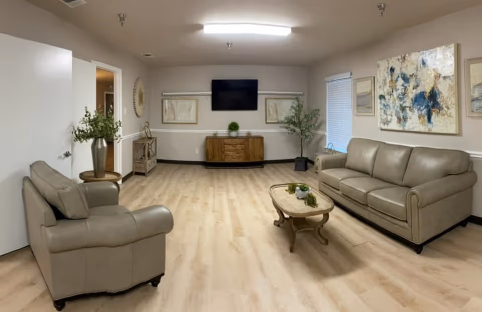 A living room with light wood flooring, beige walls, and a ceiling light. The room contains a beige leather sofa and matching armchair, a wooden coffee table with small plants, a wooden cabinet with a flat-screen TV mounted above it, and decorative framed artwork on the walls. There are also two potted plants placed near the walls.