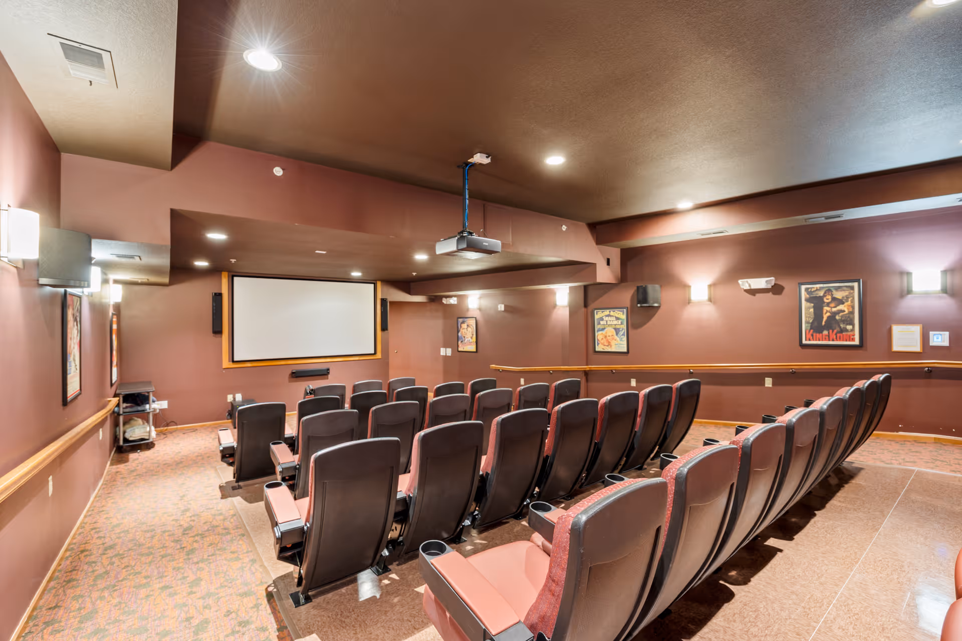 A small theater room with rows of red and black cushioned seats facing a large white projection screen. The walls are painted brown and decorated with framed movie posters. Ceiling lights illuminate the room, and a projector is mounted on the ceiling.