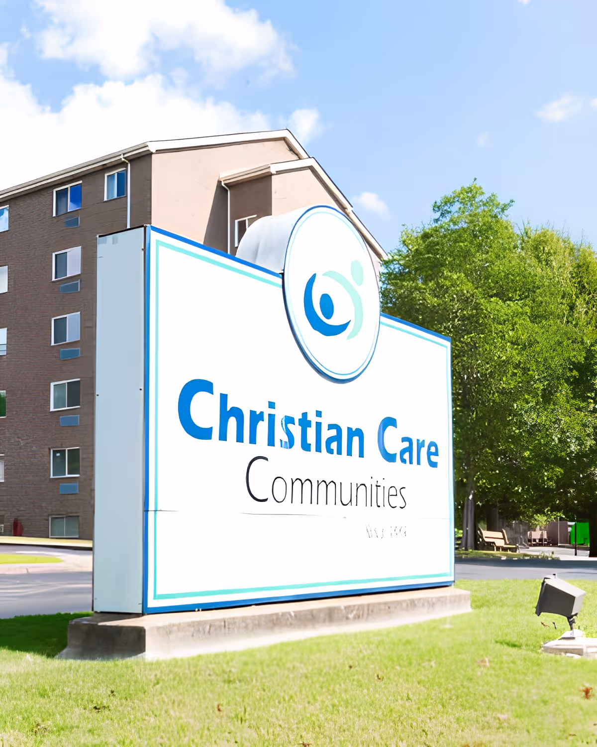 Outdoor view of a large sign for Christian Care Communities in front of a multi-story building with several windows, green grass, and trees under a partly cloudy blue sky.