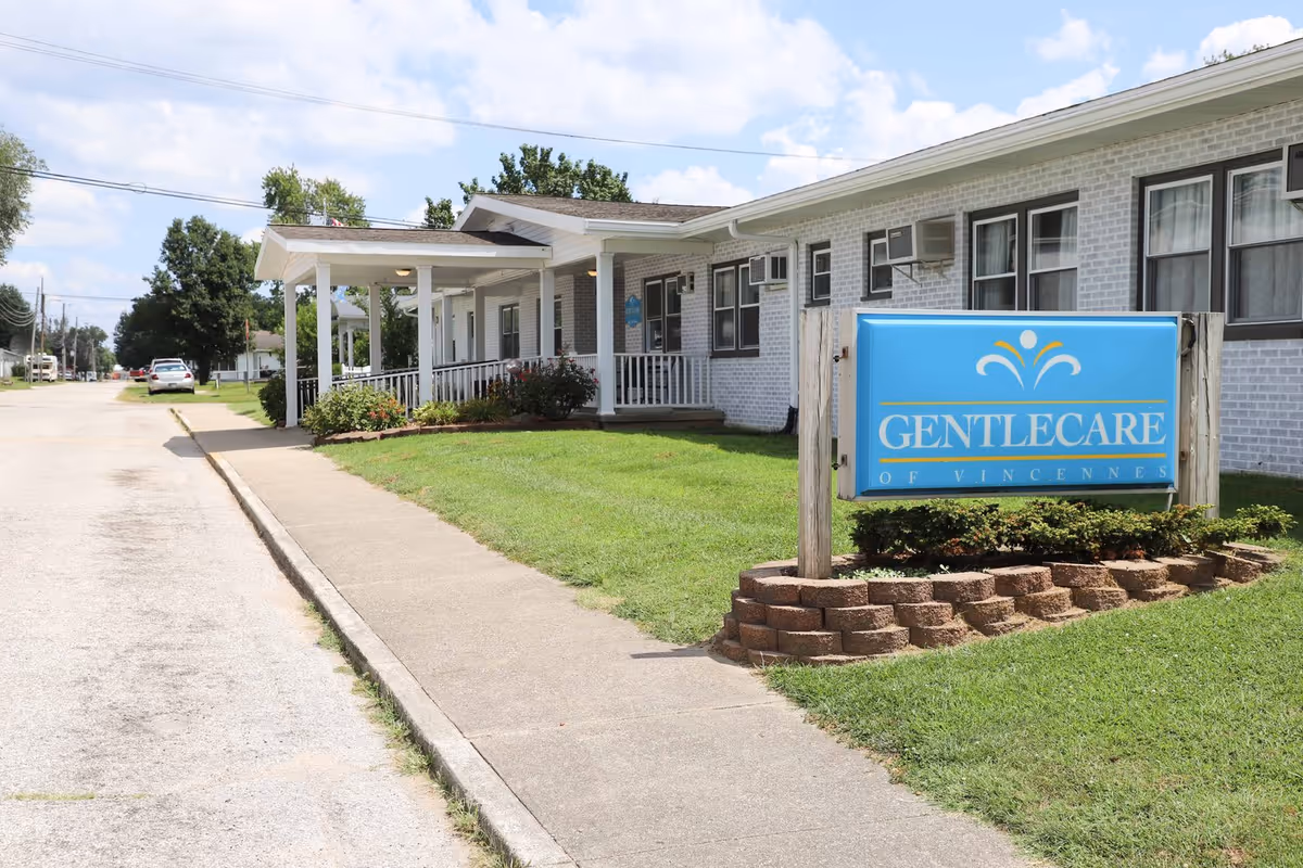 Exterior view of Gentlecare Nursing & Rehabilitation facility showing a single-story white brick building with multiple windows and air conditioning units. A blue sign with the facility's name is displayed on a landscaped area with grass and brick edging near the sidewalk. The sky is partly cloudy.