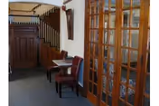 Interior view of a hallway area with a wooden staircase on the left and a small seating area with two chairs and a table. On the right side, there are wooden framed glass doors leading to another room.