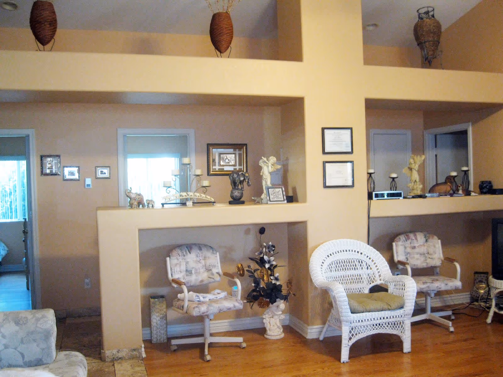 A cozy living room area with beige walls and wooden flooring. The room features a white wicker chair with a cushion, two patterned swivel chairs, and various decorative items such as candle holders, small statues, framed pictures, and a floral arrangement. There are two doorways visible leading to other rooms, and a TV is partially visible on the right side.