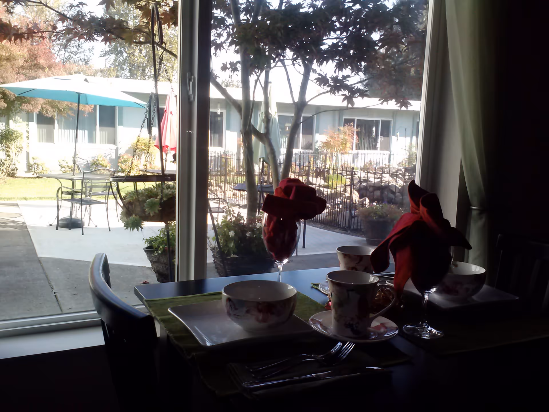 View from inside a dining room looking out through a large window to an outdoor patio area with tables, chairs, umbrellas, and trees. The dining table inside is set with cups, bowls, plates, silverware, and wine glasses with red cloth napkins folded inside.