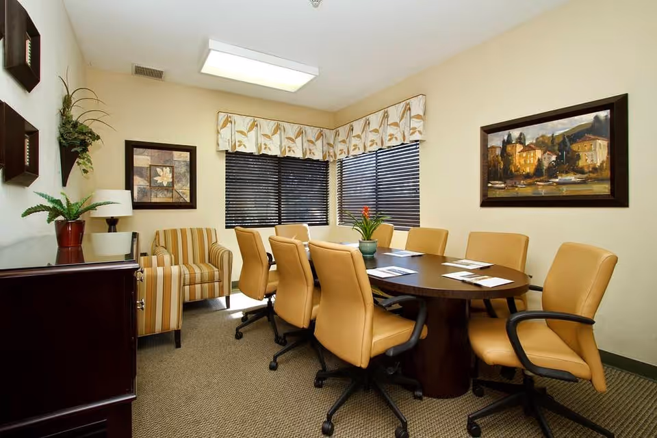 A conference room with a dark oval table surrounded by eight tan leather swivel chairs. The room has beige walls, a striped armchair in the corner, two framed paintings on the walls, a potted plant on a cabinet, and windows with black blinds and floral valances.