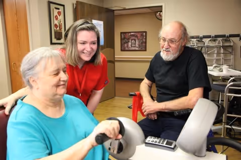 An elderly woman using a hand exercise machine while a younger woman in a red shirt smiles and supports her. An elderly man sits nearby watching. The setting appears to be a care facility room with medical equipment and framed pictures on the walls.