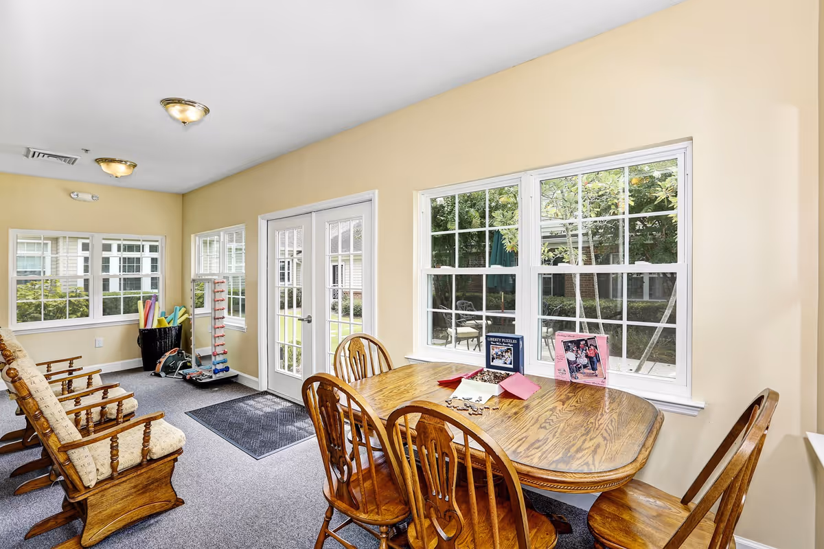 A bright room with beige walls featuring a wooden table with four chairs near windows overlooking a garden. On the table are two puzzle boxes and some puzzle pieces. To the left, there are three cushioned wooden rocking chairs and a basket with colorful pool noodles and exercise equipment. French doors lead outside to a patio area with chairs and greenery.