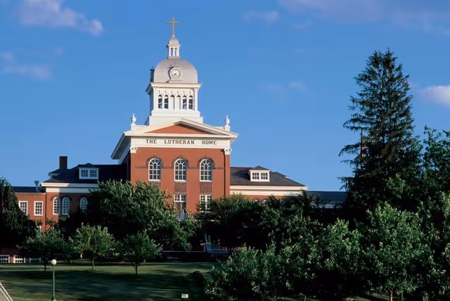 Red brick building with a clock tower and cross, surrounded by green trees and blue sky.