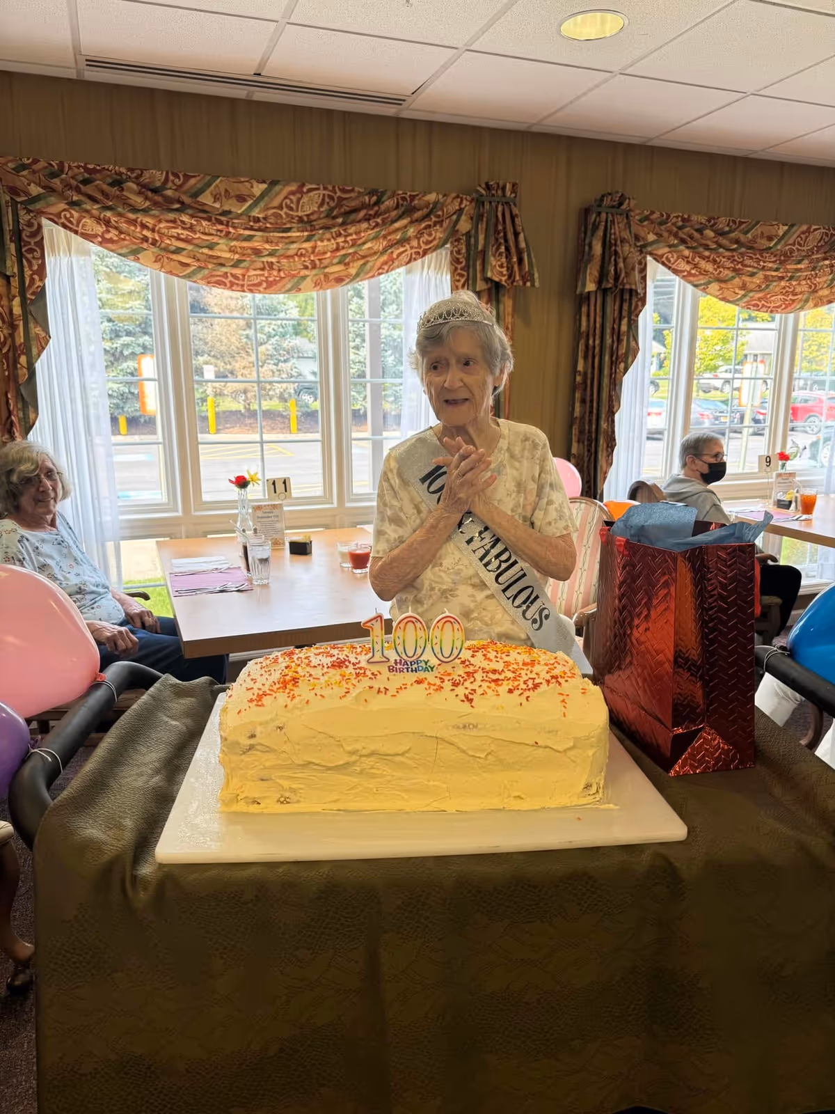 An elderly woman wearing a tiara and sash stands behind a large rectangular cake with a '100' topper in a dining room.