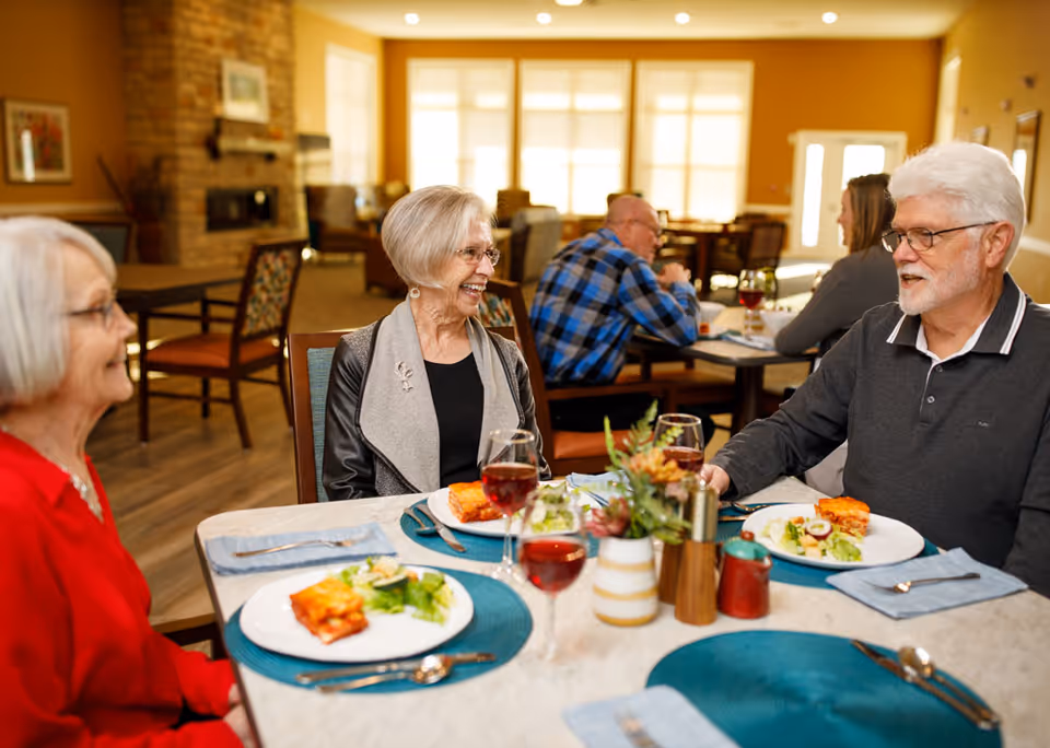 Elderly residents sit around a table in a bright dining room enjoying a meal and conversation.