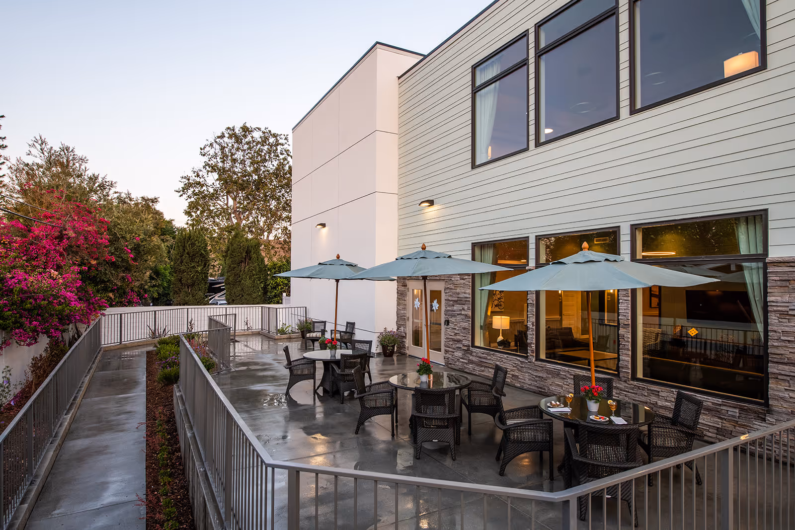 Outdoor patio with round tables, umbrellas and wicker chairs beside a two-story memory care building.