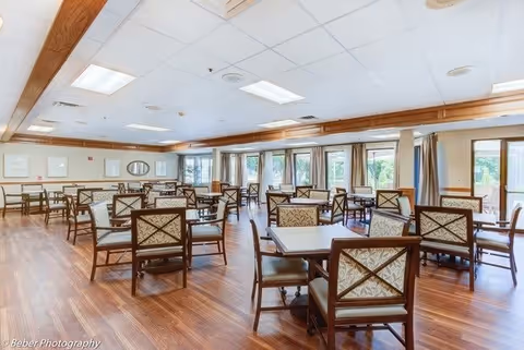 A spacious dining room with multiple square tables and cushioned chairs arranged neatly on a wooden floor. Large windows with curtains allow natural light to fill the room, and the ceiling has recessed lighting panels. The room has a warm and inviting atmosphere.