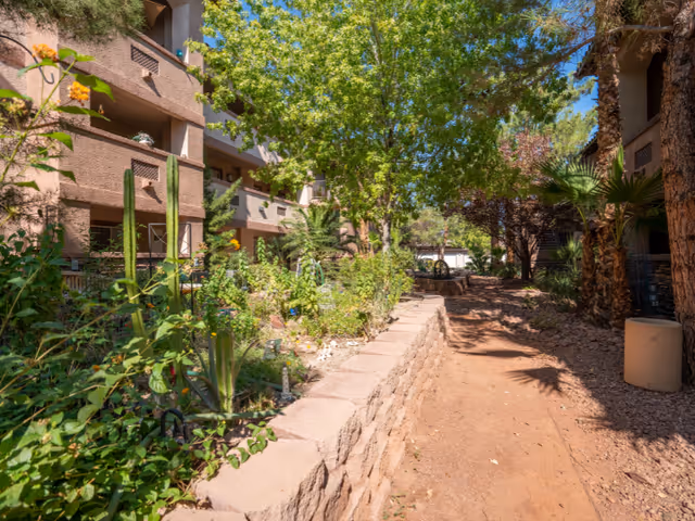 Outdoor garden area at a senior living facility with a dirt pathway, stone retaining wall, various green plants including cacti and trees, and brown multi-story buildings on either side under a clear blue sky.