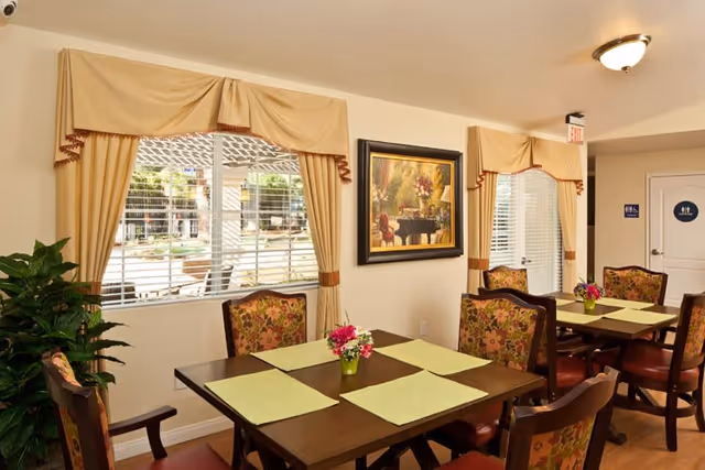 A dining area with two wooden tables, each set with four green placemats and small flower arrangements in the center. The chairs have floral patterned upholstery. There are two windows with beige curtains and valances, and a framed painting on the wall. A door with restroom signs is visible in the background.