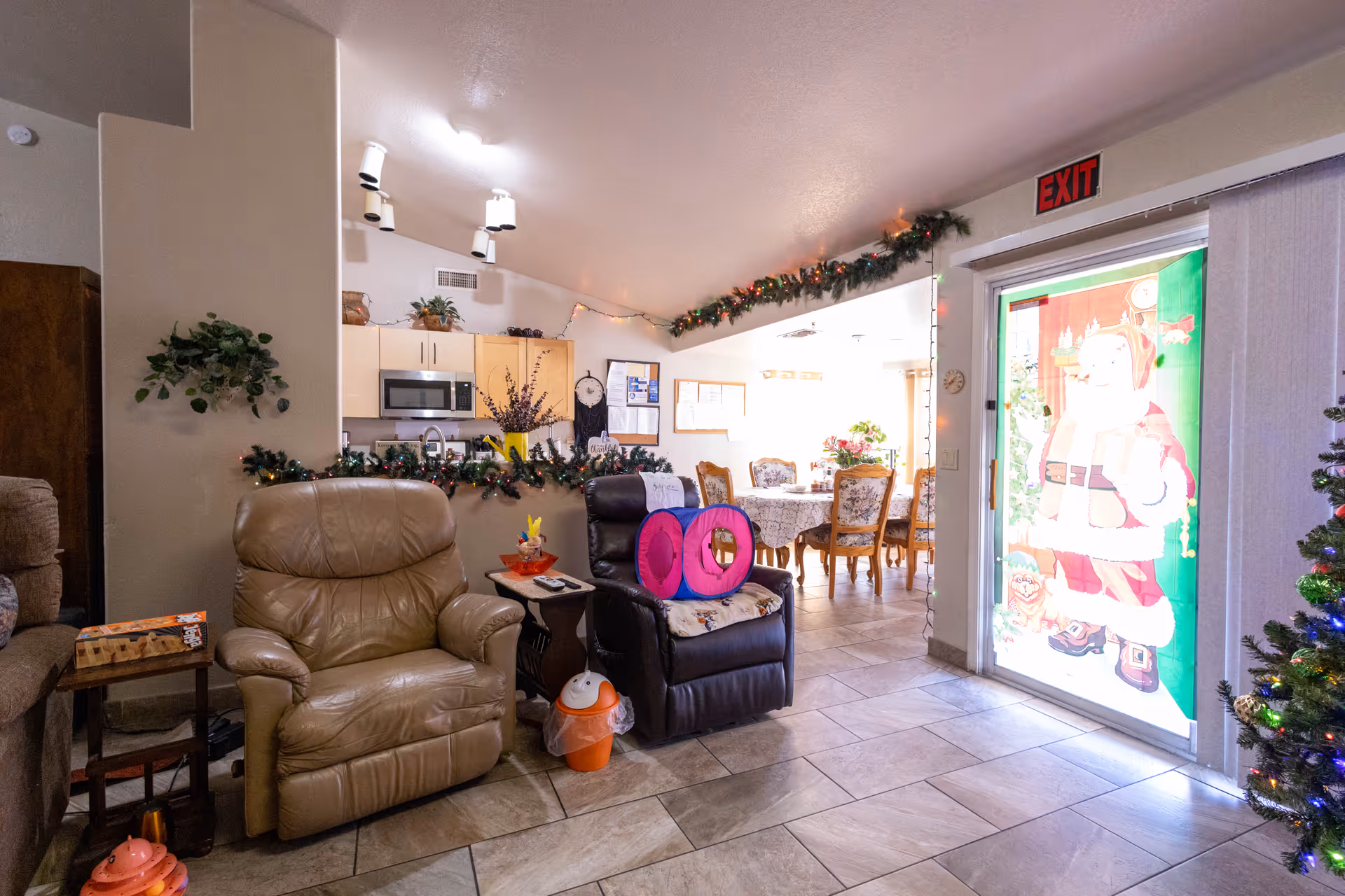 Interior common living area with recliner chairs, holiday garlands, an open kitchen and dining area, and a glass door decorated with a Santa image.