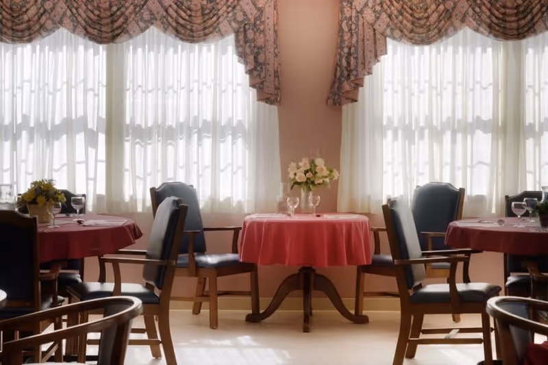 A dining room with round tables covered in red tablecloths, each set with glassware and floral centerpieces. The room has large windows with sheer white curtains and decorative valances, allowing natural light to fill the space. Wooden chairs with dark cushions surround the tables.