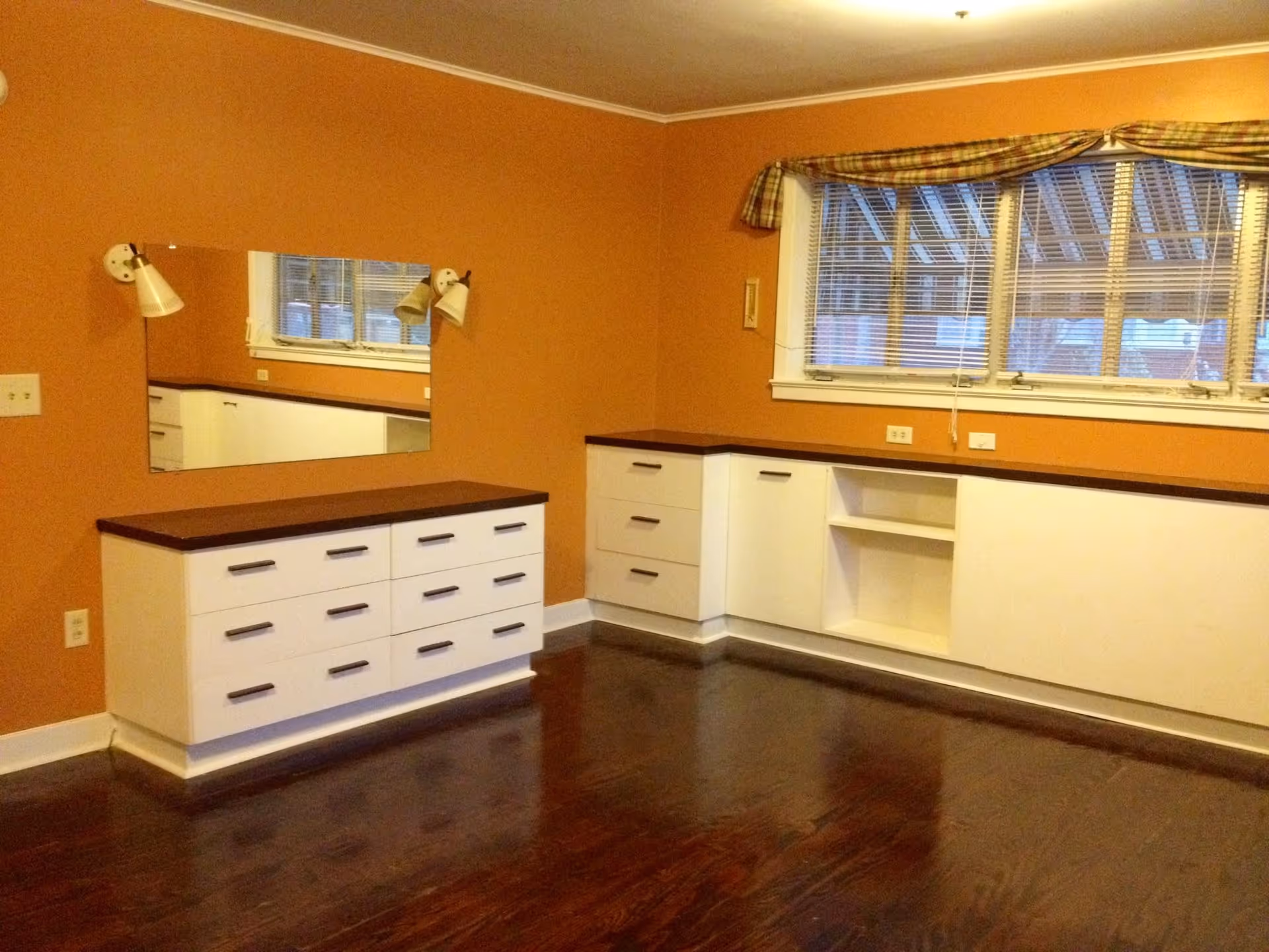 An empty room with orange walls and dark wooden floor. The room features white built-in cabinets with dark brown countertops along two walls, a rectangular mirror mounted on one wall, two wall-mounted lamps on either side of the mirror, and a window with blinds and a plaid valance.