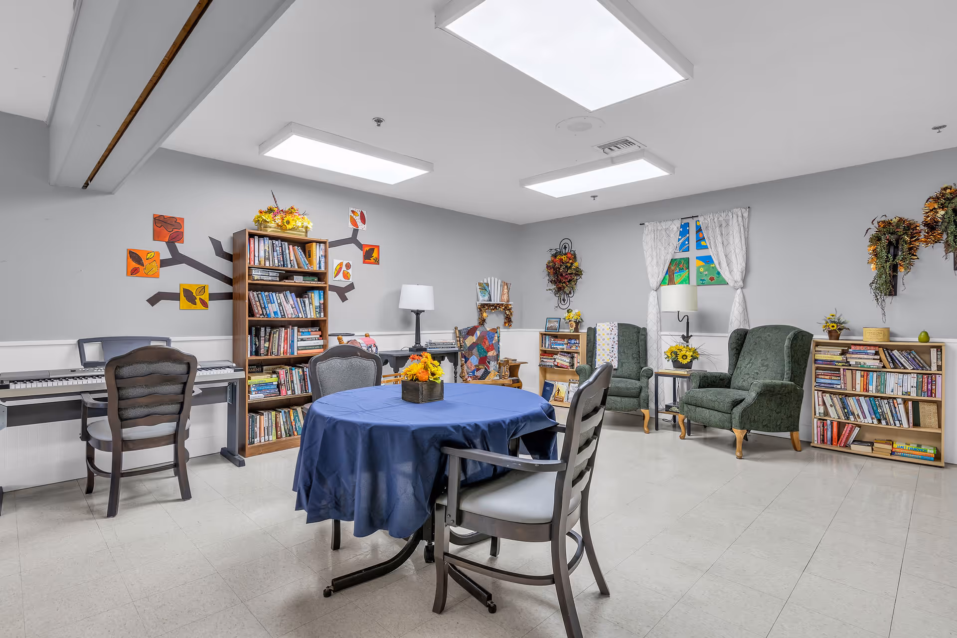 A cozy common room with a round table covered with a blue tablecloth and four chairs around it. The room has bookshelves filled with books, two green upholstered armchairs, a keyboard piano with a chair, and decorative wall art including a tree with colorful leaves and wreaths. The walls are light gray with white trim, and the floor is light-colored tile.