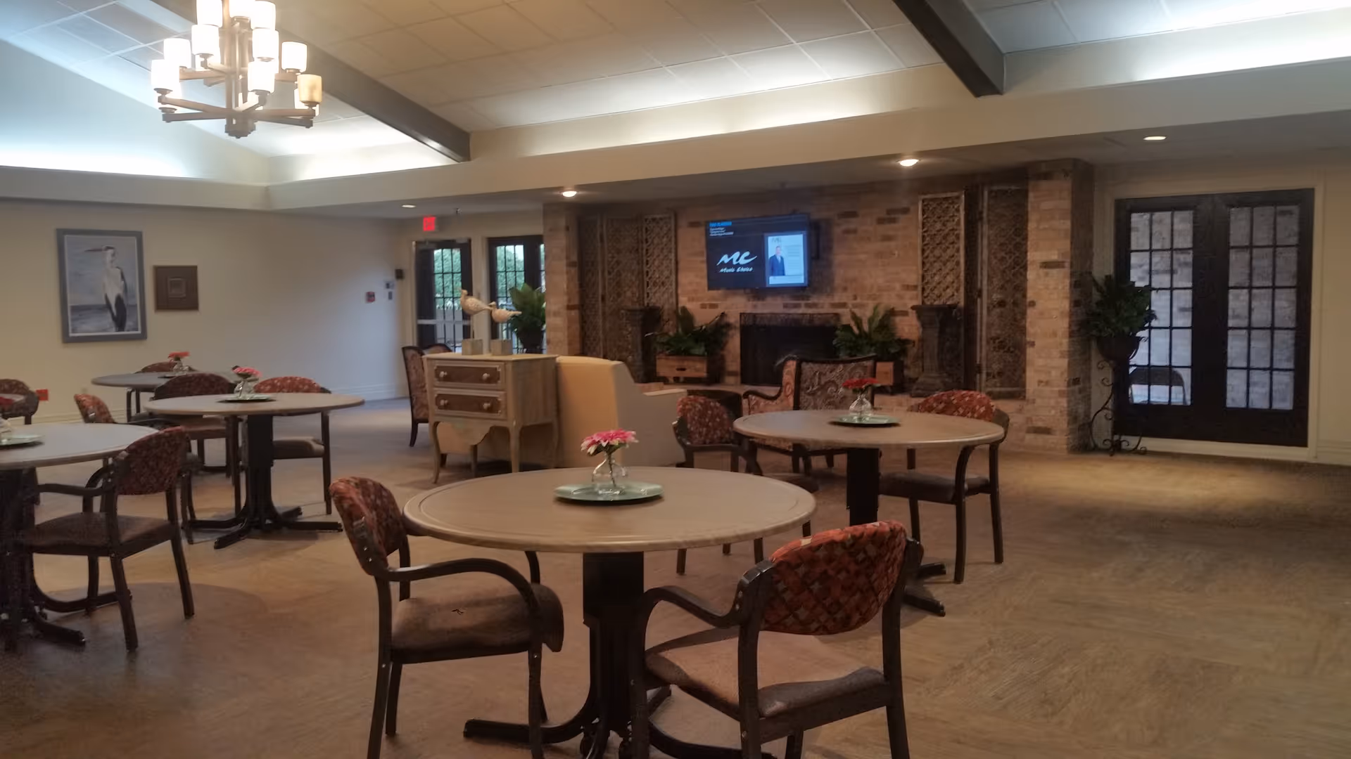 A spacious common area with several round tables and cushioned chairs arranged around them. Each table has a small flower centerpiece. The room features a brick wall with a mounted flat-screen TV above a fireplace, flanked by decorative wooden panels and plants. There are double glass doors on the right side and a chandelier hanging from the ceiling.