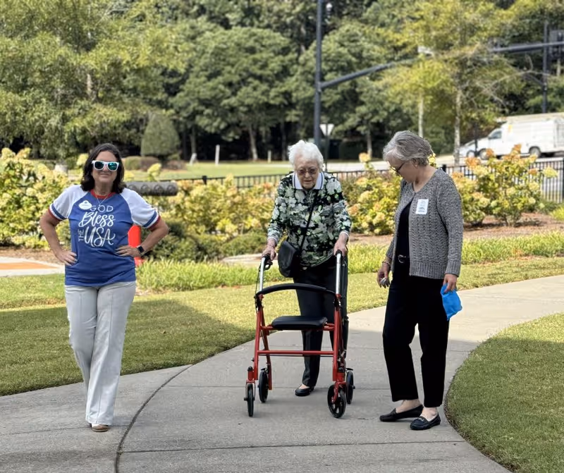 Three women walking outside on a paved pathway surrounded by greenery. One elderly woman uses a red walker, accompanied by two other women, one wearing a 'God Bless the USA' shirt and sunglasses, and the other in a gray cardigan holding a blue mask.