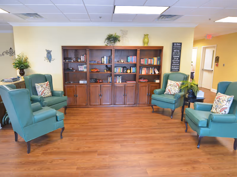 A cozy common area with four green upholstered armchairs arranged around a wooden floor. Behind the chairs is a large wooden bookshelf filled with books, decorative items, and plants. The walls are painted light yellow, and there are additional plants and wall decor. A hallway with doors is visible in the background.