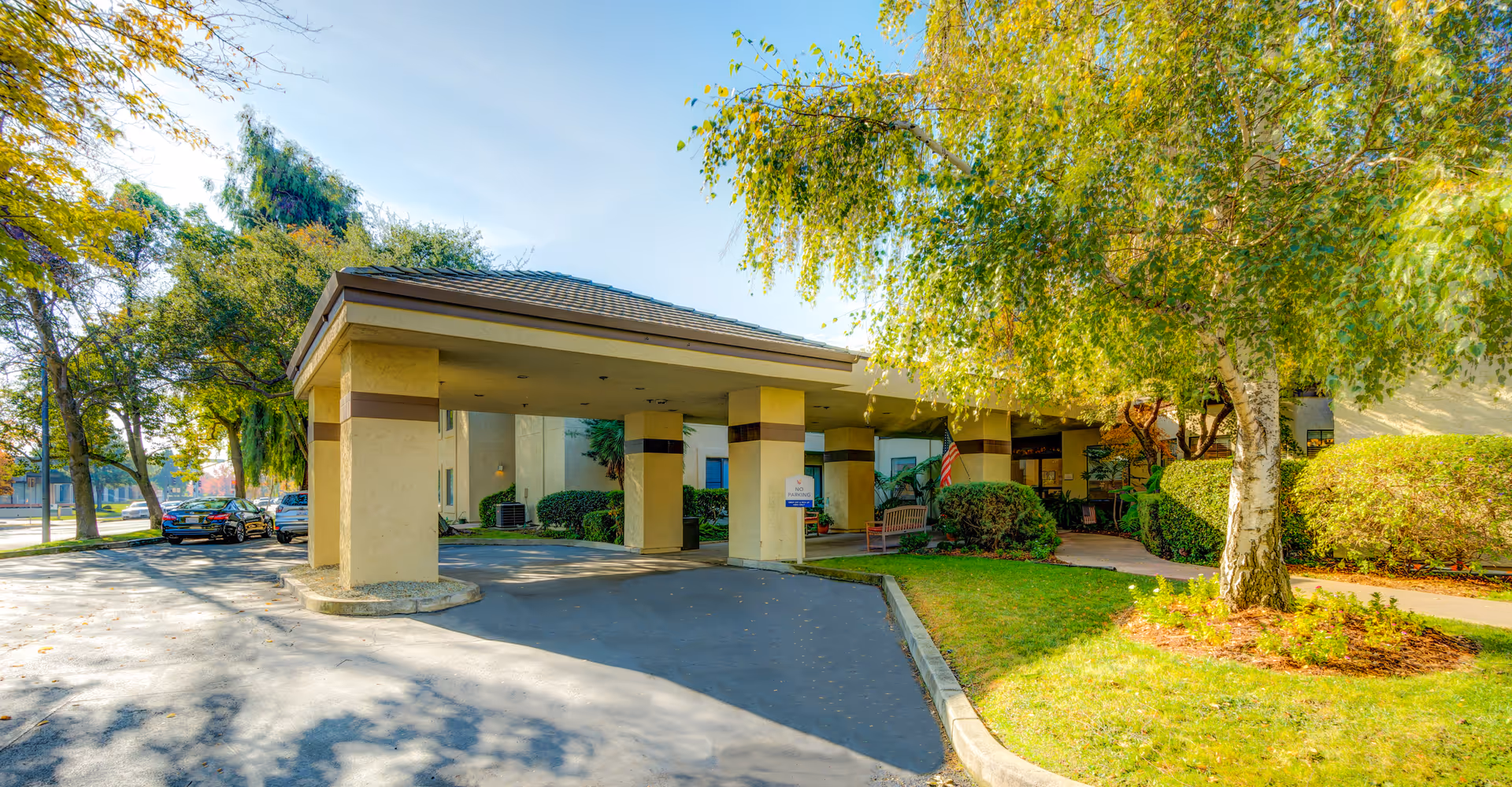 Covered entrance canopy and drive-up area of a senior living building surrounded by trees, parked cars, and landscaped greenery.