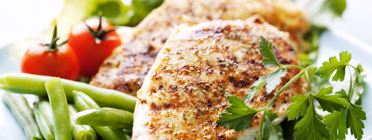 Close-up of a plate with grilled chicken breast seasoned with herbs, green beans, cherry tomatoes, and a sprig of fresh parsley.
