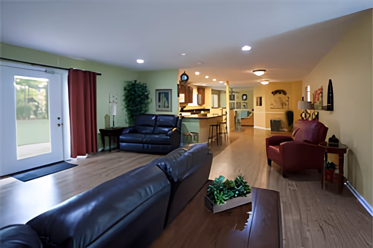 A spacious living room area with leather sofas and a red armchair. The room has wooden flooring, light green and yellow walls, recessed ceiling lights, and a glass door with red curtains leading outside. There is a kitchen area visible in the background with bar stools and a counter.
