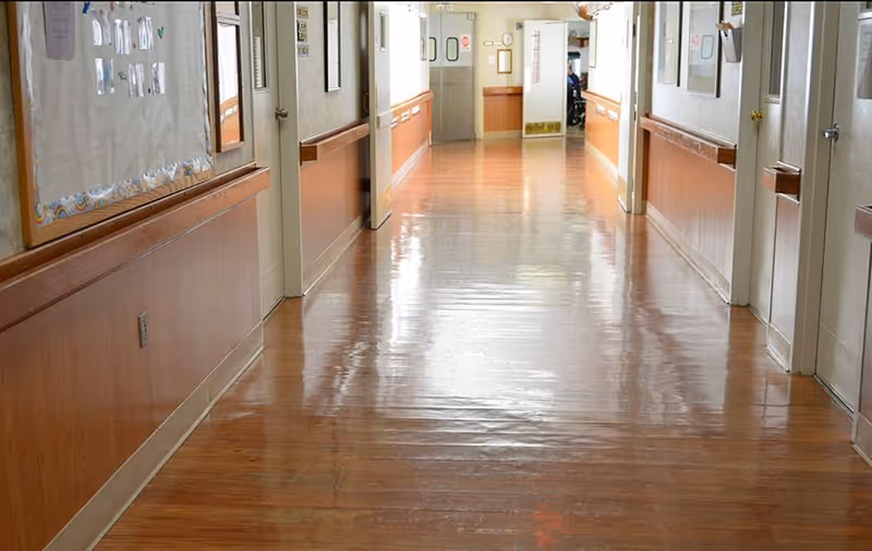 A long, clean hallway with polished wooden floors and wood-paneled lower walls in a senior living facility. Doors and bulletin boards line both sides of the corridor, with a double door at the far end.