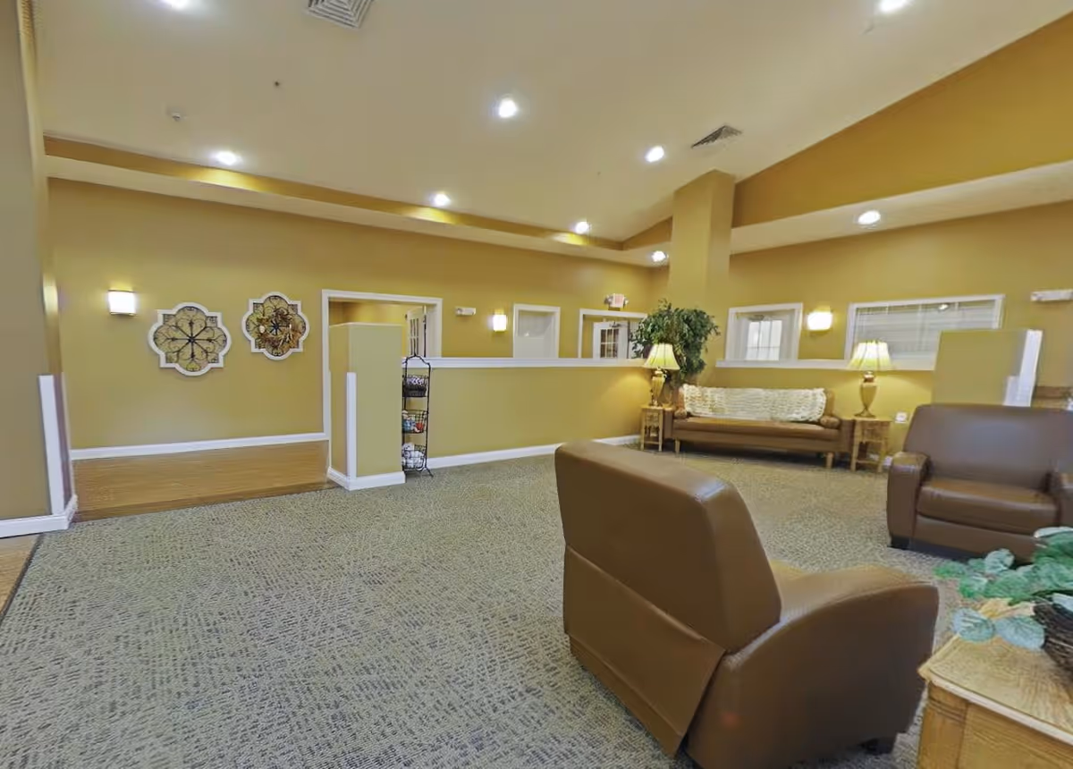 A spacious and well-lit common area with beige walls and carpeted floor. The room features brown leather chairs, a wooden sofa with a white throw blanket, side tables with lamps, decorative wall art, and a potted plant. The ceiling has recessed lighting and air vents.