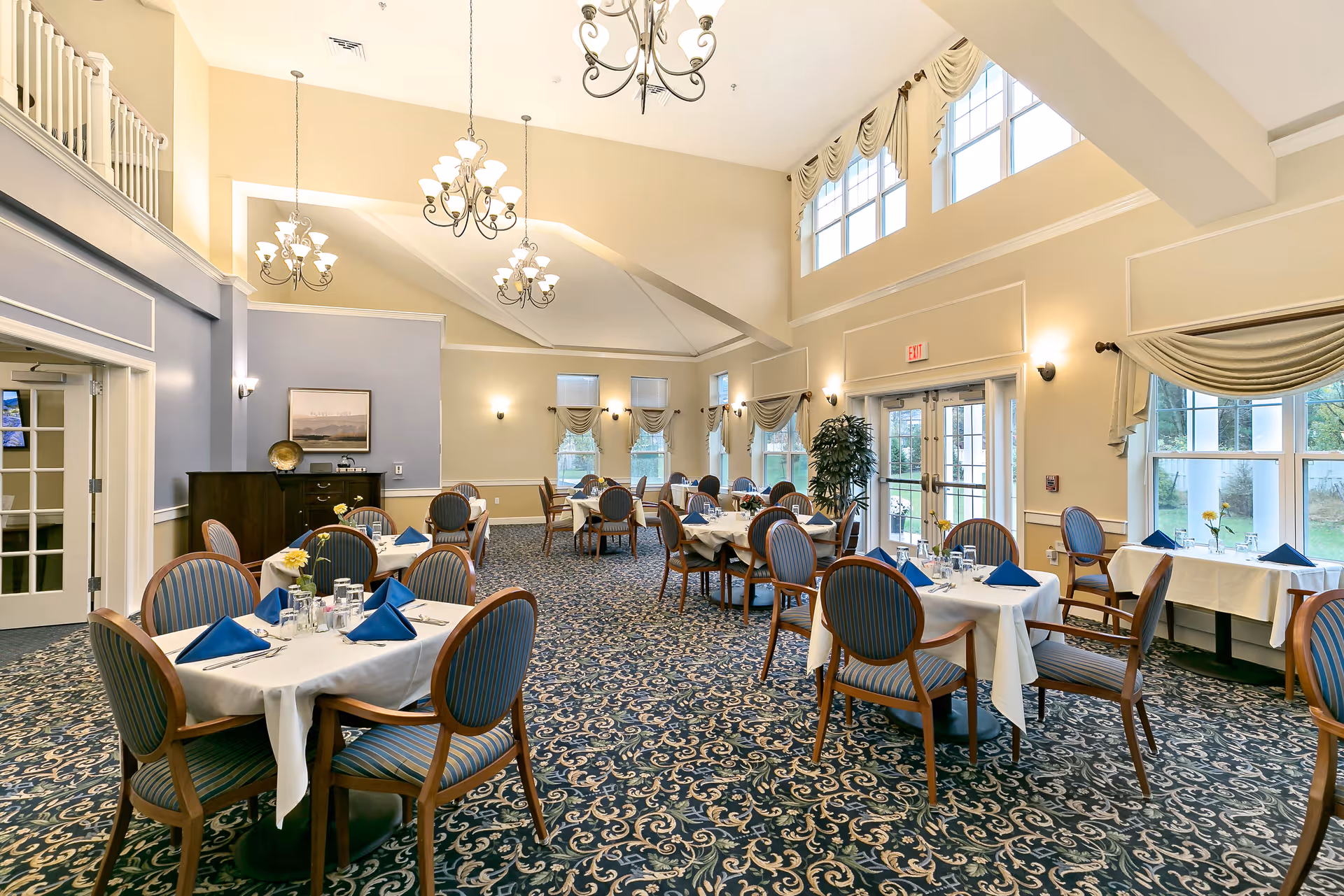 Spacious, elegant dining room with round tables set with white tablecloths and blue napkins under chandeliers.