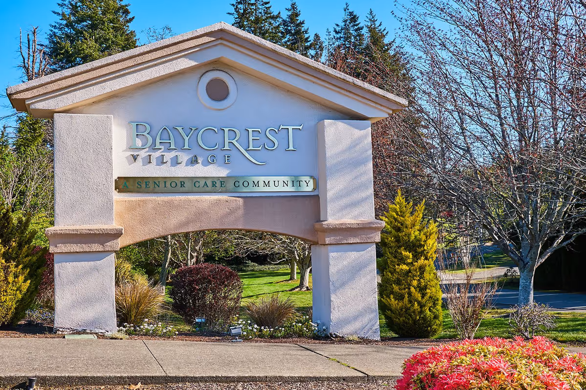 Entrance sign for Baycrest Village, a senior care community, surrounded by landscaped greenery including bushes, trees, and colorful plants under a clear blue sky.