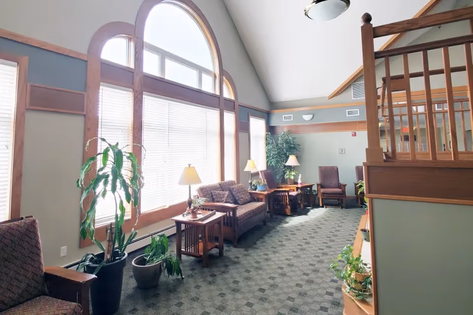 A bright and spacious senior living facility common area with large arched windows letting in natural light. The room features comfortable seating including armchairs and a loveseat, wooden side tables with lamps, and several potted plants. The walls are painted in soft tones with wooden trim, and the ceiling is vaulted with a ceiling light fixture. A wooden railing is visible on the right side.