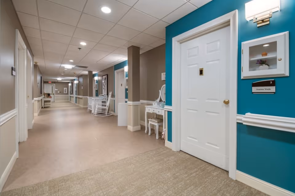 Well-lit interior hallway of a senior living facility with doors, rocking chairs, and a teal accent wall.