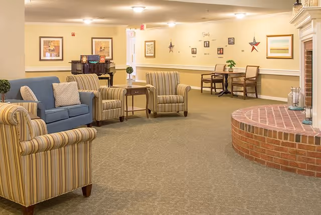 A cozy senior living community lounge area with striped armchairs, a blue sofa with cushions, a small wooden table, a brick fireplace, and a piano in the background. The walls are decorated with framed pictures and wall art, and there is a small dining table with chairs in the back.