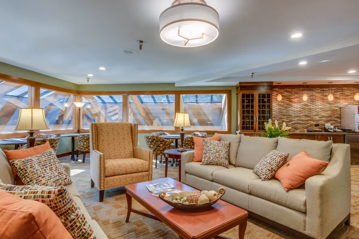 Comfortable communal living room with sofas and armchairs around a coffee table, large windows with angled wooden beams, and a kitchenette/bar in the background.