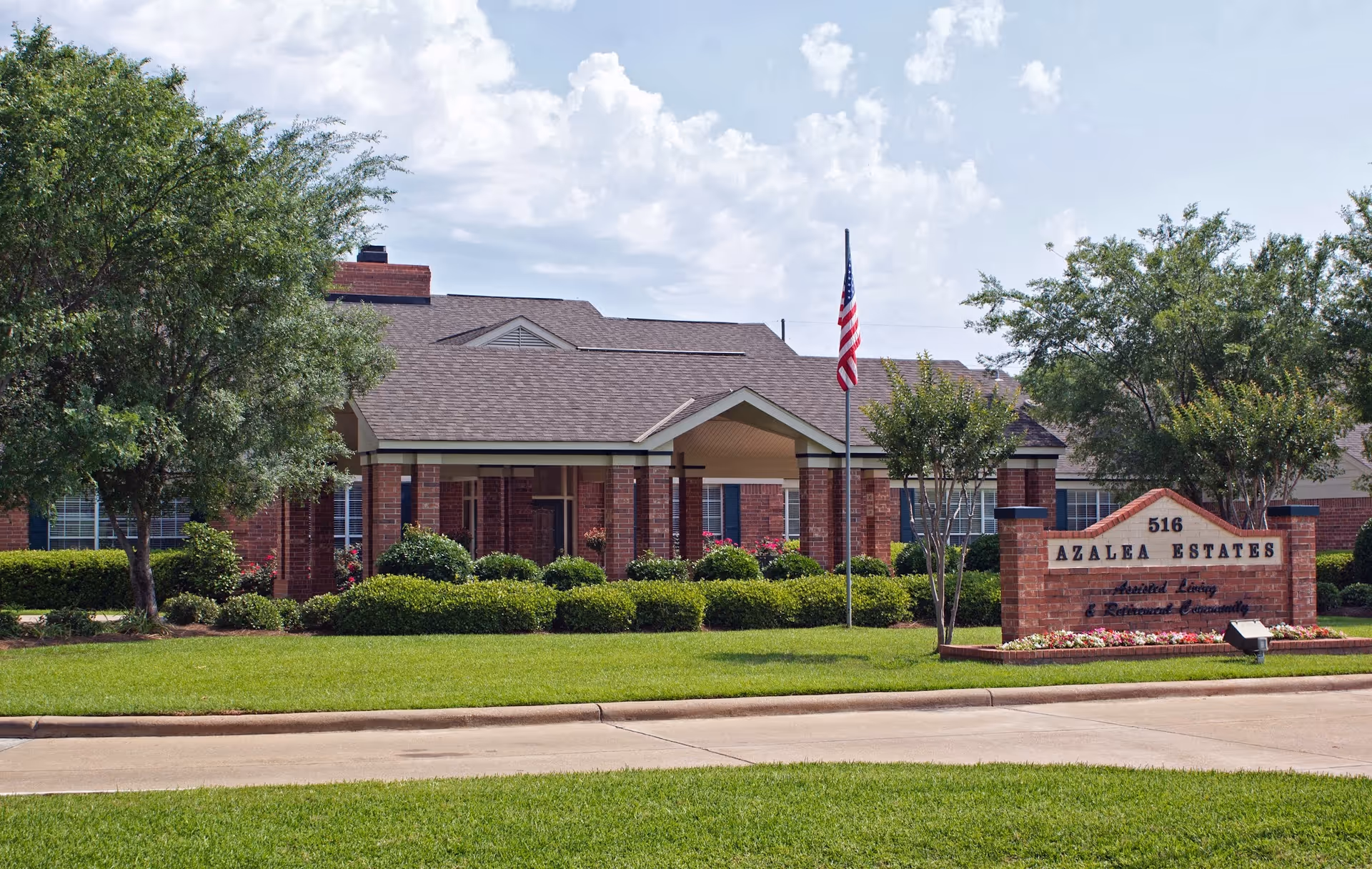 Exterior view of Azalea Estates senior living facility showing a single-story brick building with a covered entrance, well-maintained green lawn, bushes, trees, an American flag on a flagpole, and a brick sign with the facility name and address.