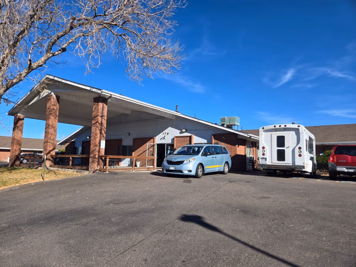 Entrance of a single-story brick senior living building with a covered porte-cochère, parked vehicles, and a clear blue sky.