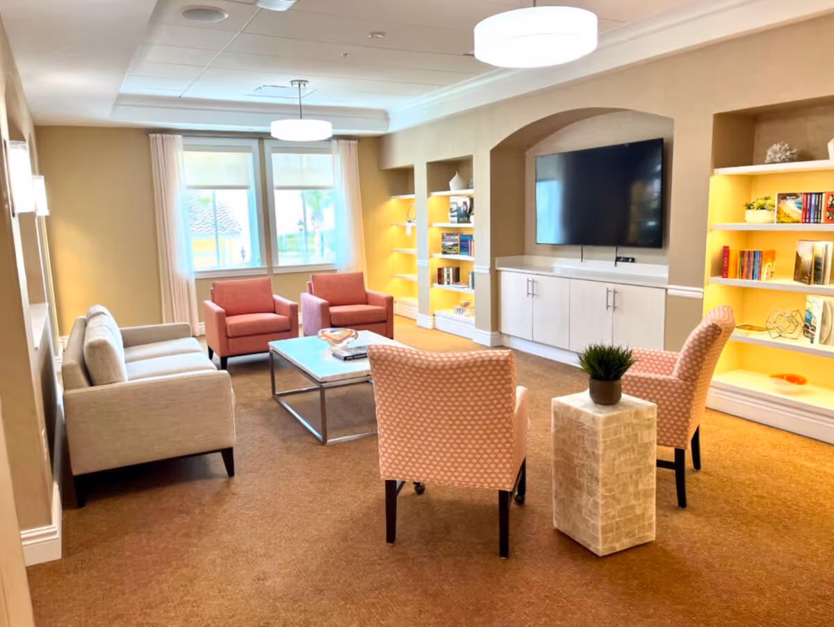A cozy living room with beige walls and carpet, featuring a beige sofa, two pink armchairs, a white coffee table, and a large flat-screen TV mounted on the wall. Built-in shelves with books and decorative items are illuminated with warm lighting. Two large windows with white curtains allow natural light into the room.