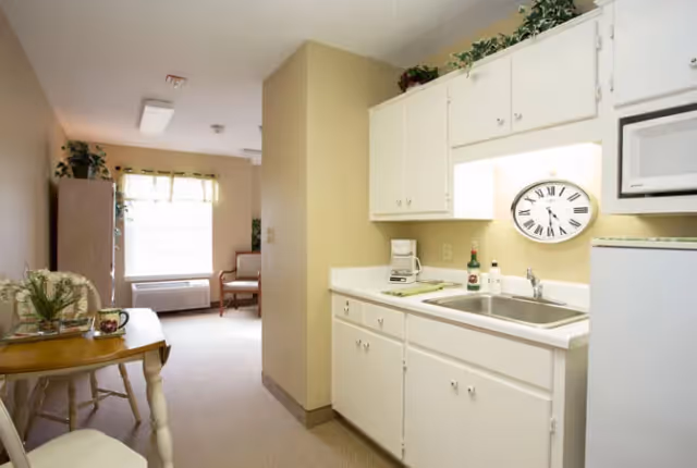 Small kitchenette with white cabinets and sink opening into a bright sitting area with a table and chairs.