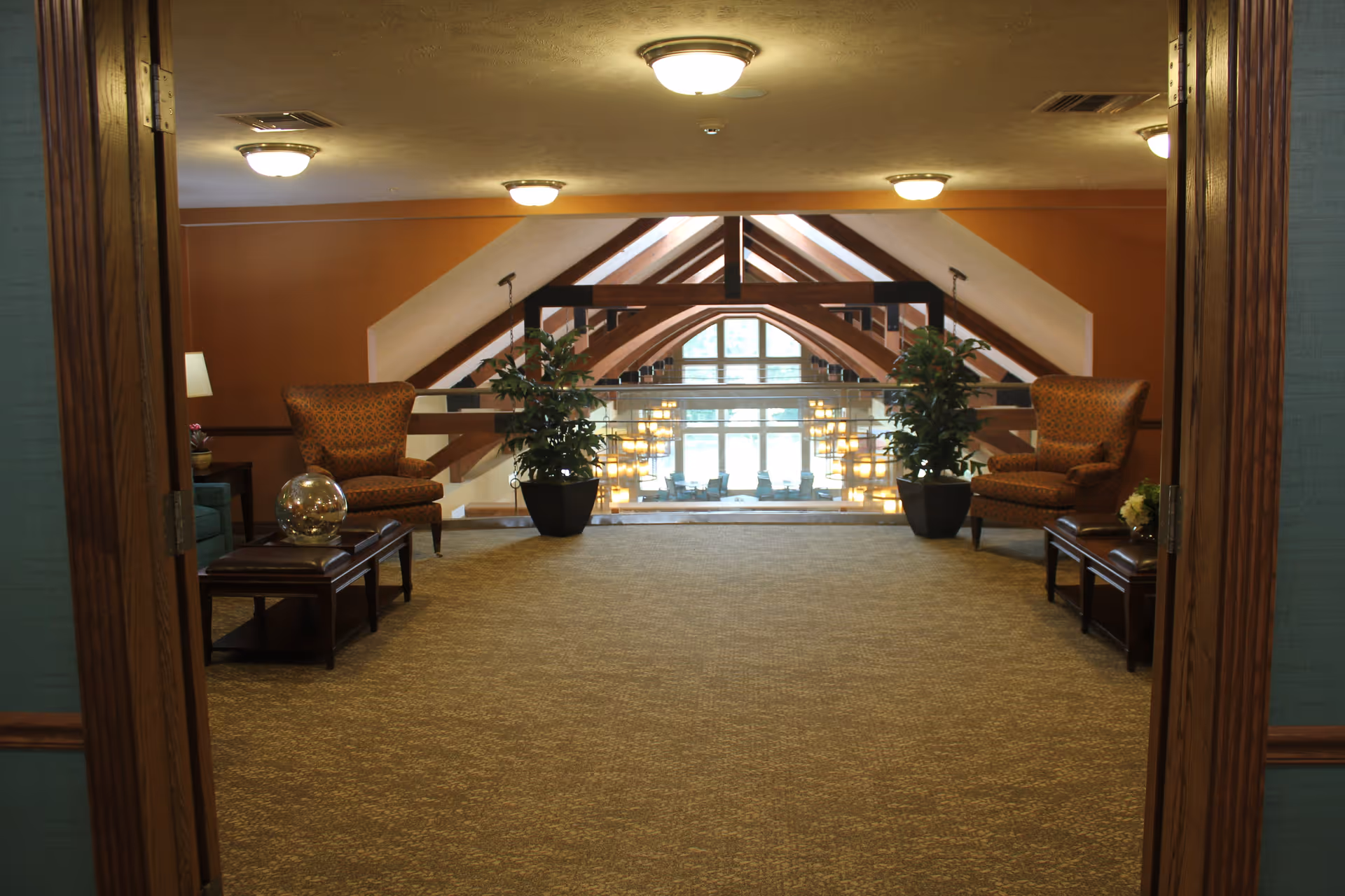 Interior view of a seating area in a retirement community with two patterned armchairs, two small tables, potted plants, and a large window with wooden beams and hanging lights in the background.