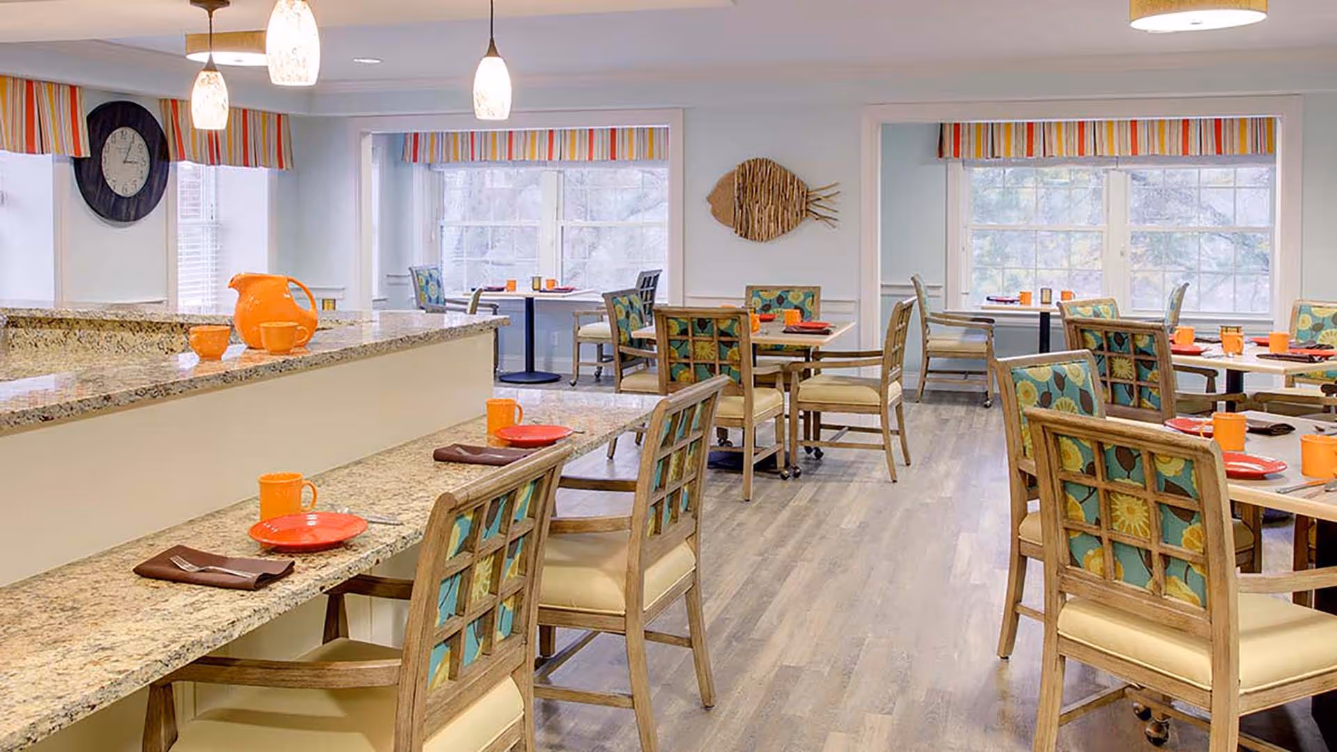 A bright dining room with multiple tables and chairs, each table set with orange cups and red plates. The chairs have wooden frames with colorful floral upholstery. The room features large windows with striped valances, pendant lights hanging from the ceiling, and a granite countertop with additional seating. A decorative fish wall art is visible on the far wall.