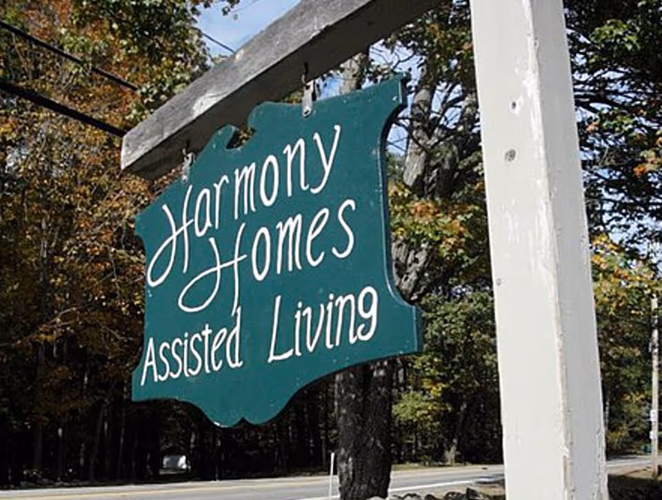A green wooden hanging sign with white cursive and print text that reads 'Harmony Homes Assisted Living' mounted on a white post with trees and a road in the background.