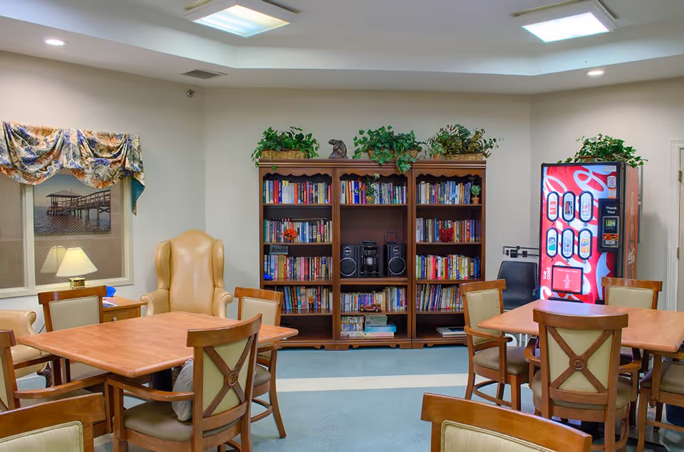 A cozy common area with wooden tables and chairs arranged for group seating. There is a large wooden bookshelf filled with books and a stereo system in the center. A vending machine is visible in the corner, and a window with floral curtains and a lamp on a side table is on the left side of the room. The room has soft lighting from ceiling fixtures and plants on top of the bookshelf and vending machine.