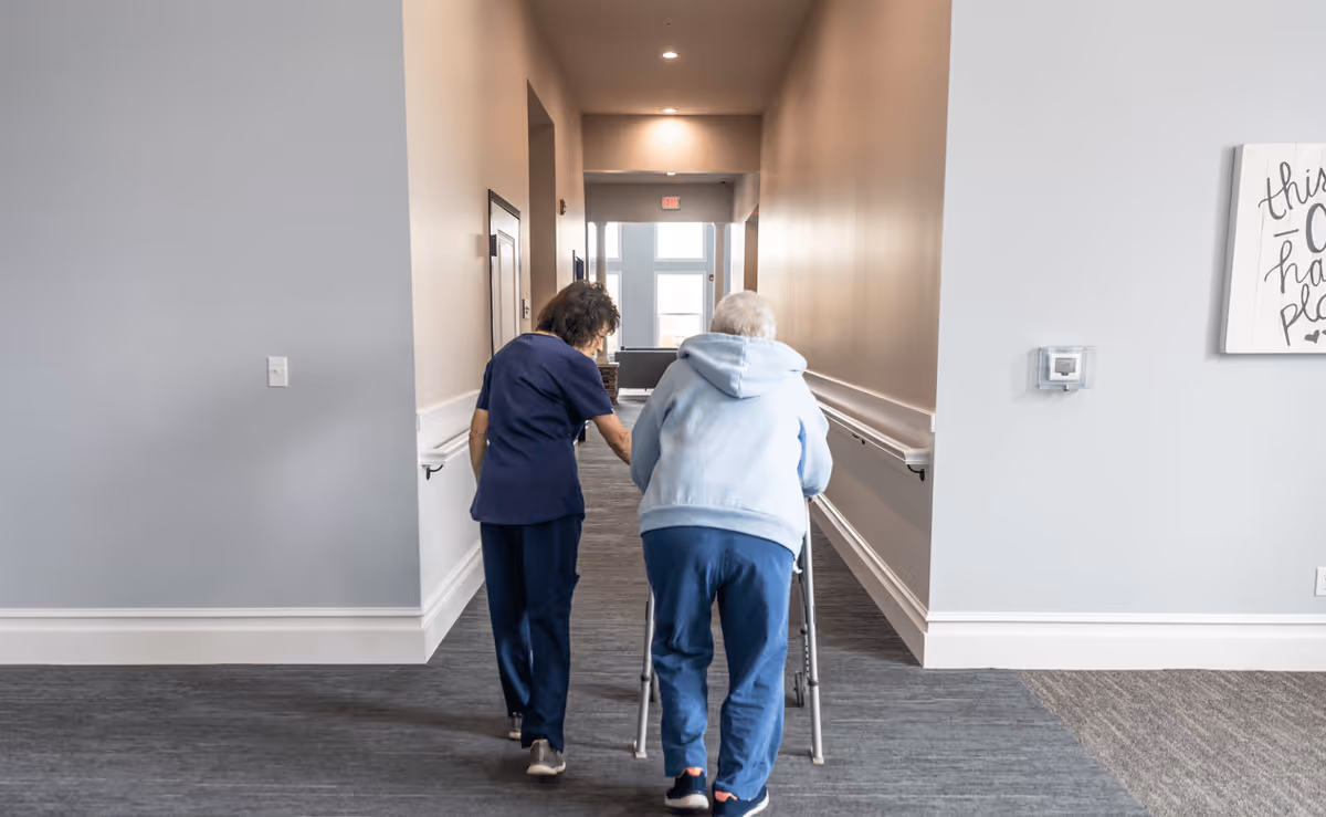 A caregiver assisting an elderly woman using a walker as they walk down a well-lit hallway with handrails on both sides in a senior living facility.