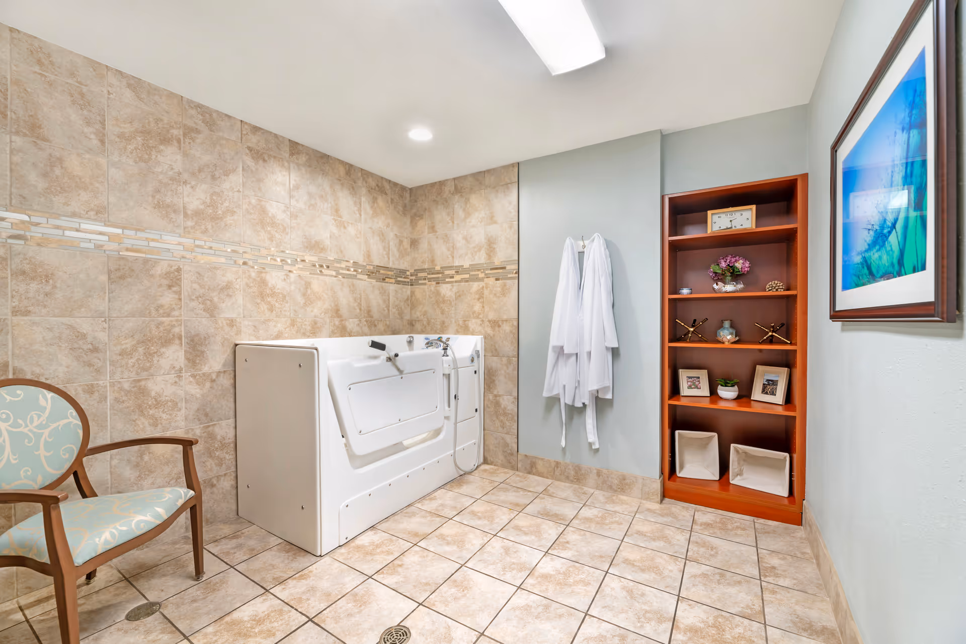 Tiled bathing room with a walk-in bathtub, robes on hooks, a shelving unit with decor, and a chair.