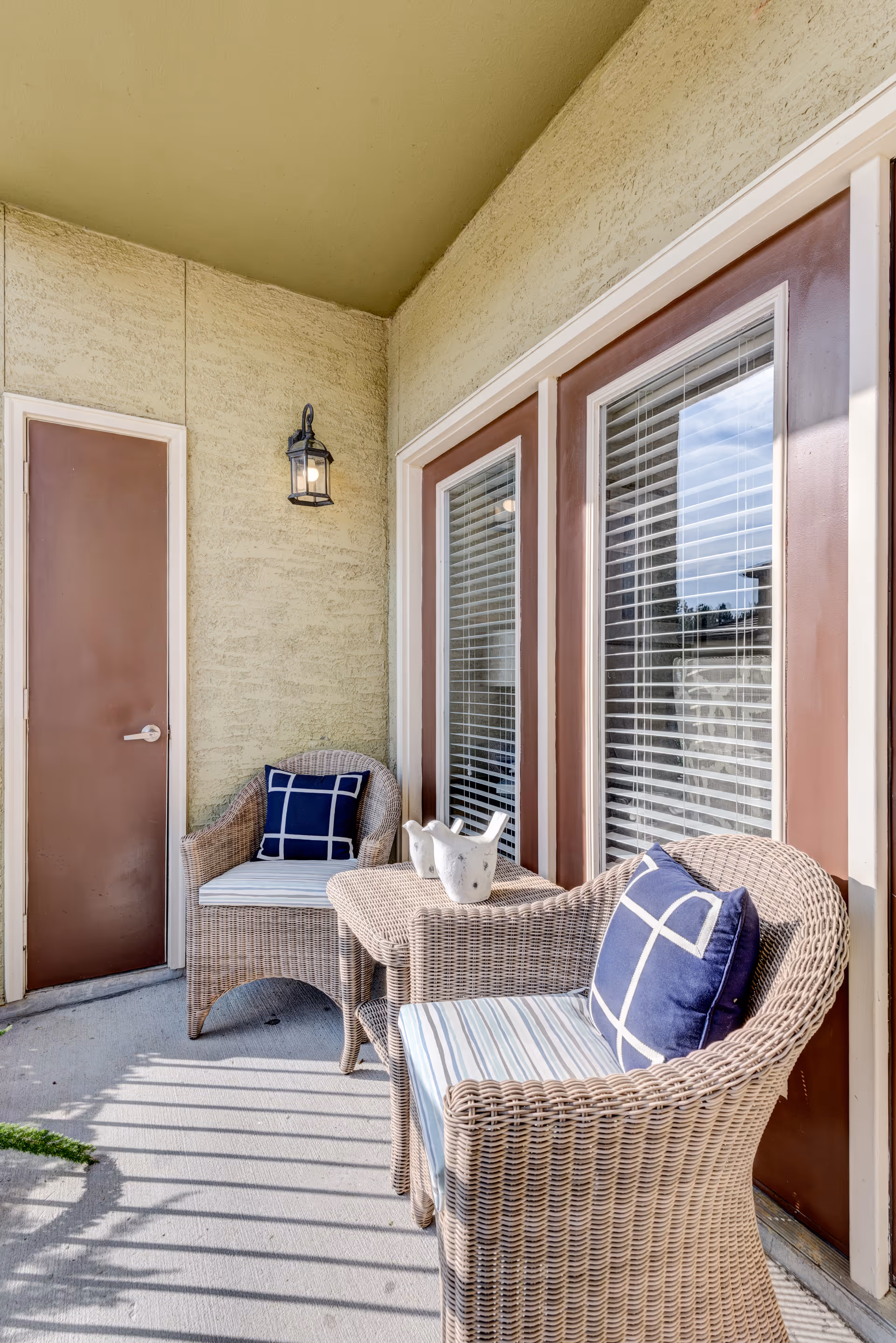 A small outdoor patio area with two wicker chairs featuring blue and white patterned cushions, a matching wicker table between them with a decorative white ceramic bird, a brown door, and a wall-mounted lantern light fixture.
