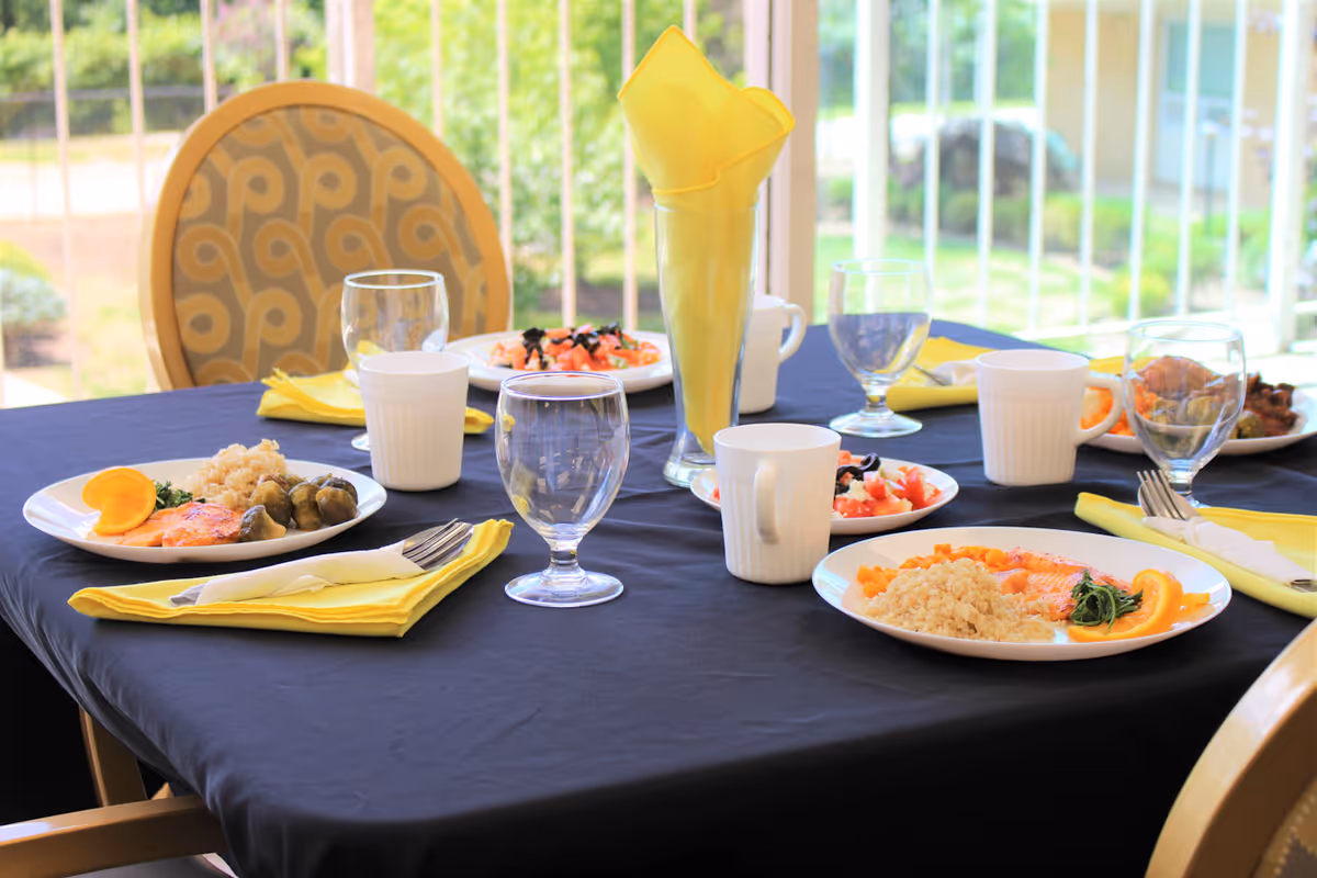 A dining table set with plates of food including rice, vegetables, and orange slices, along with glasses, cups, and yellow napkins. The table is covered with a dark blue tablecloth and is situated near a window with a view of greenery outside.