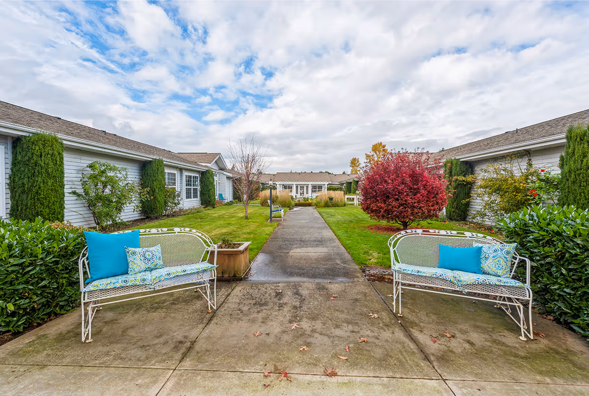 Courtyard with a central walkway flanked by decorative metal benches, shrubs, and single-story senior living buildings under a cloudy sky.