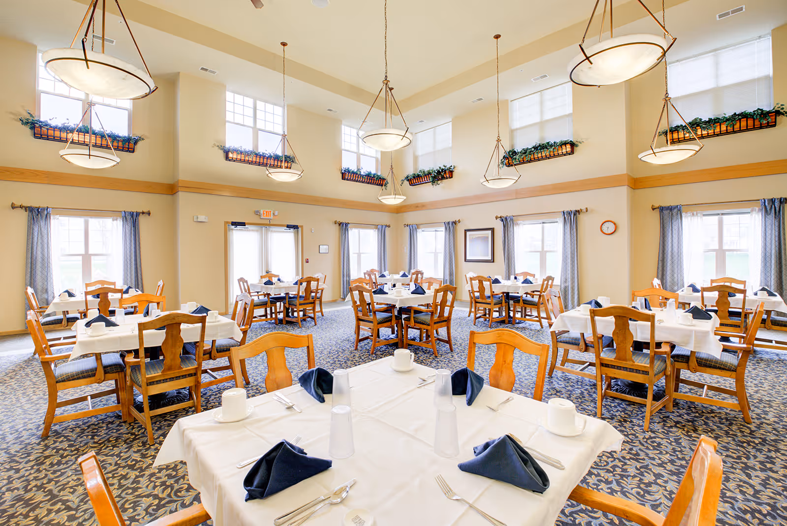 A bright and spacious dining room with multiple wooden tables and chairs arranged neatly. Each table is covered with a white tablecloth and set with navy blue folded napkins, cups, and utensils. Large windows with blue curtains allow natural light to fill the room, and hanging pendant lights provide additional illumination. The walls are beige with decorative plants placed on ledges near the ceiling.