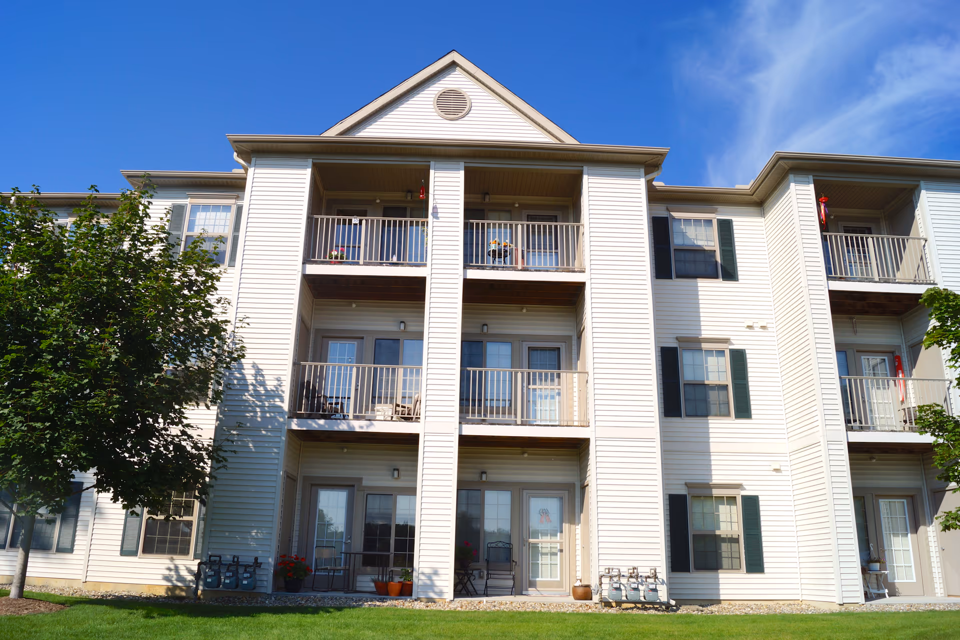Three-story beige siding apartment building with balconies, green shutters, trees and a lawn under a blue sky.